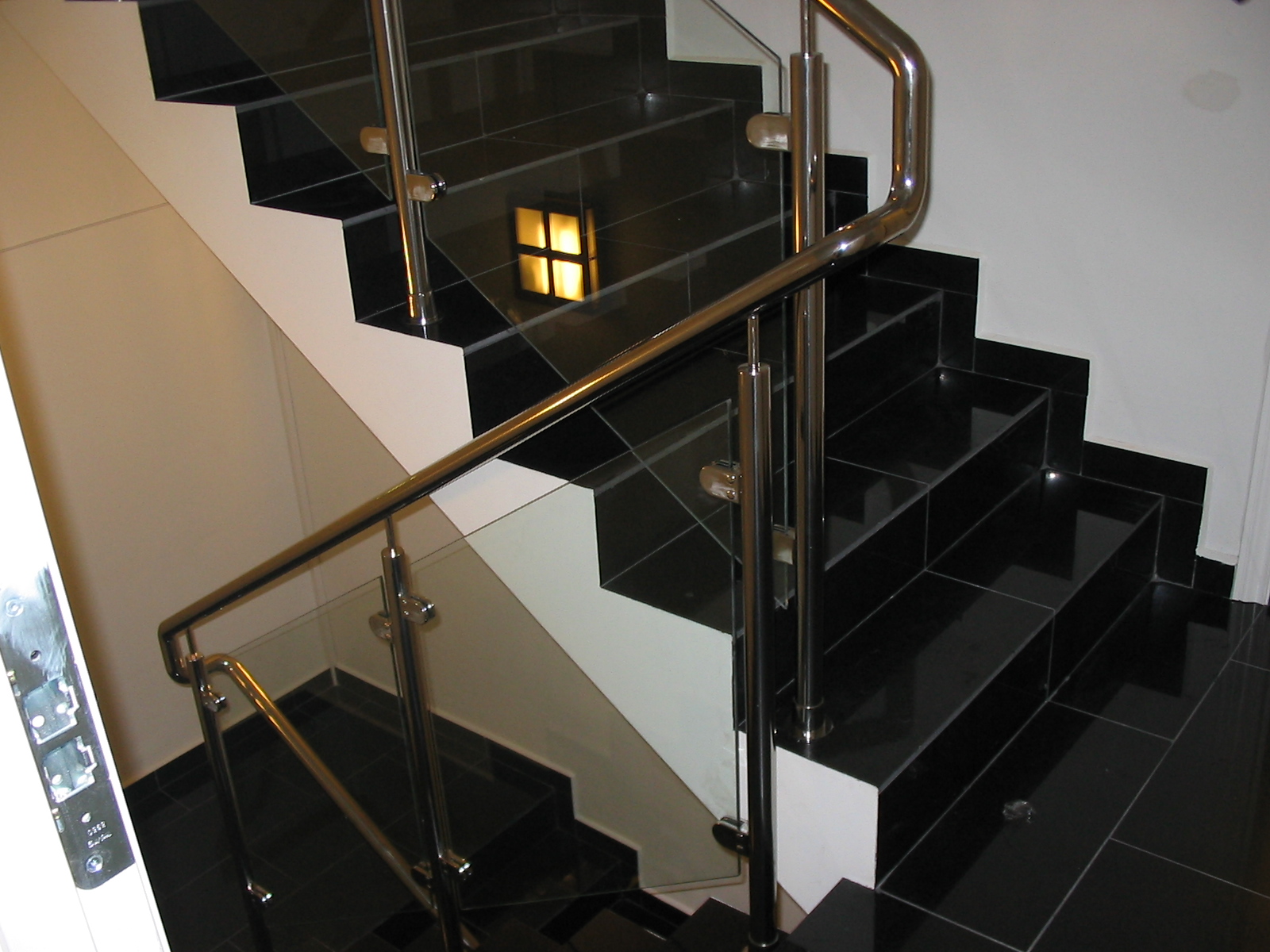 Czarne, błyszczące schody wyłożone płytkami z balustradą ze stali nierdzewnej i szkła, widok z dołu, w tle odbicie światła.